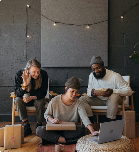 Group of three people sitting on the floor and chairs, looking at a laptop in a cozy room with string lights
